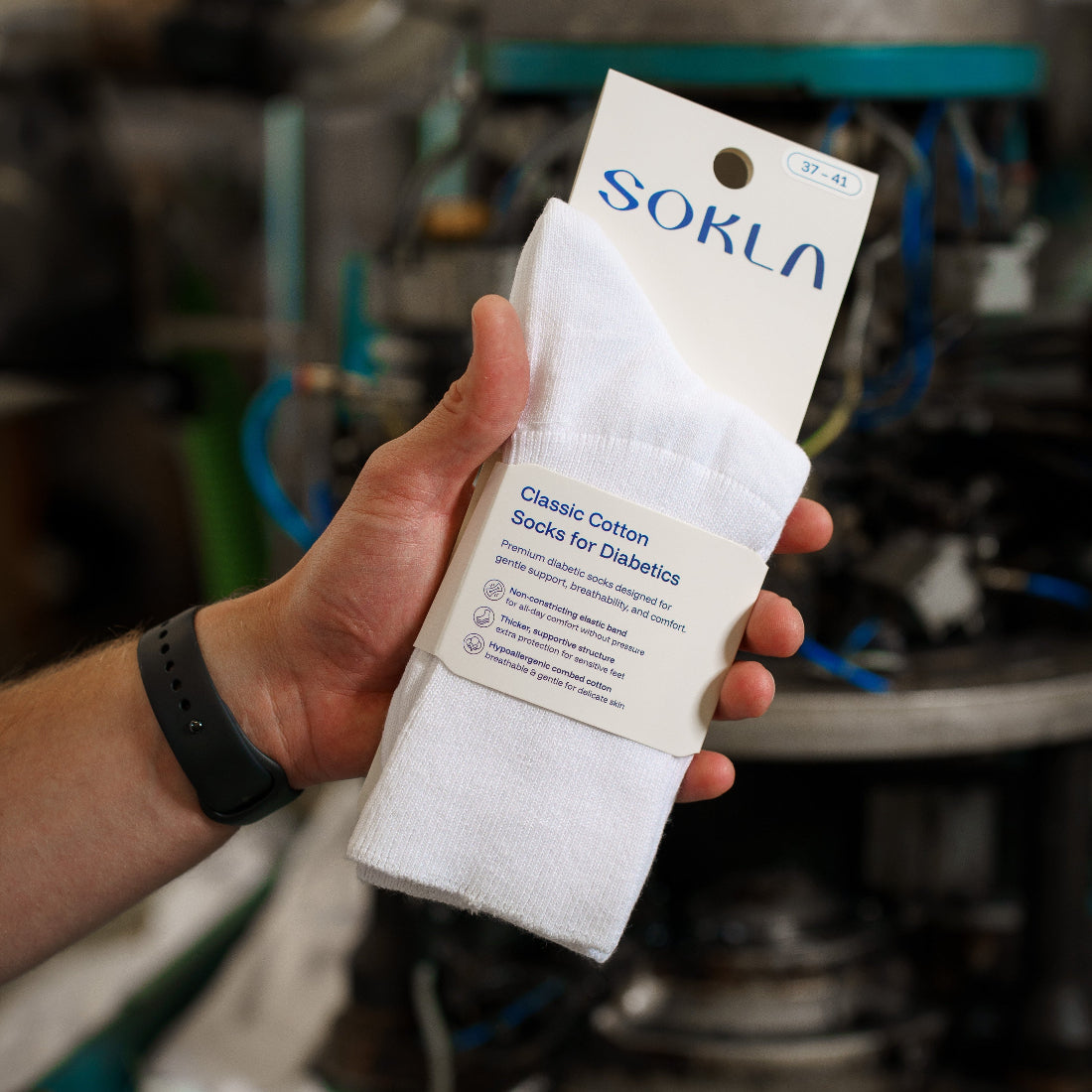 A hand holds a packaged pair of white socks in front of an industrial machine. The packaging card shows the brand name SOKLA and reads Classic Cotton socks for Diabetics
